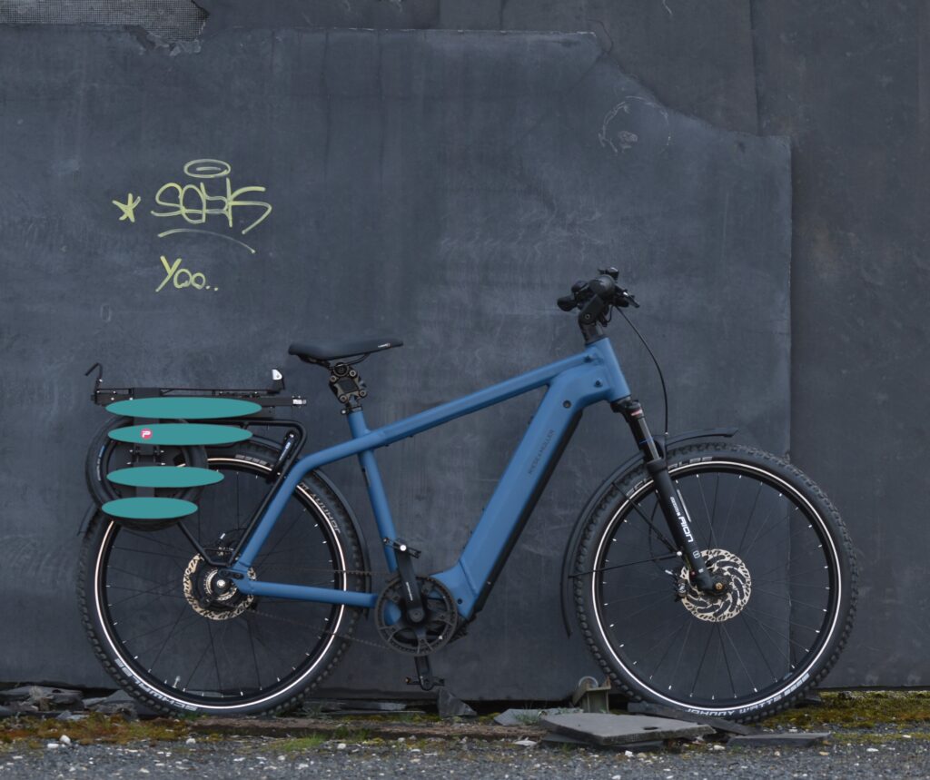 Blaues Riese & Müller Fahrrad mit türkisem, eingeklappten Pullit Fahrradanhänger auf dem Gepäckträger vor grauer Schieferwand.