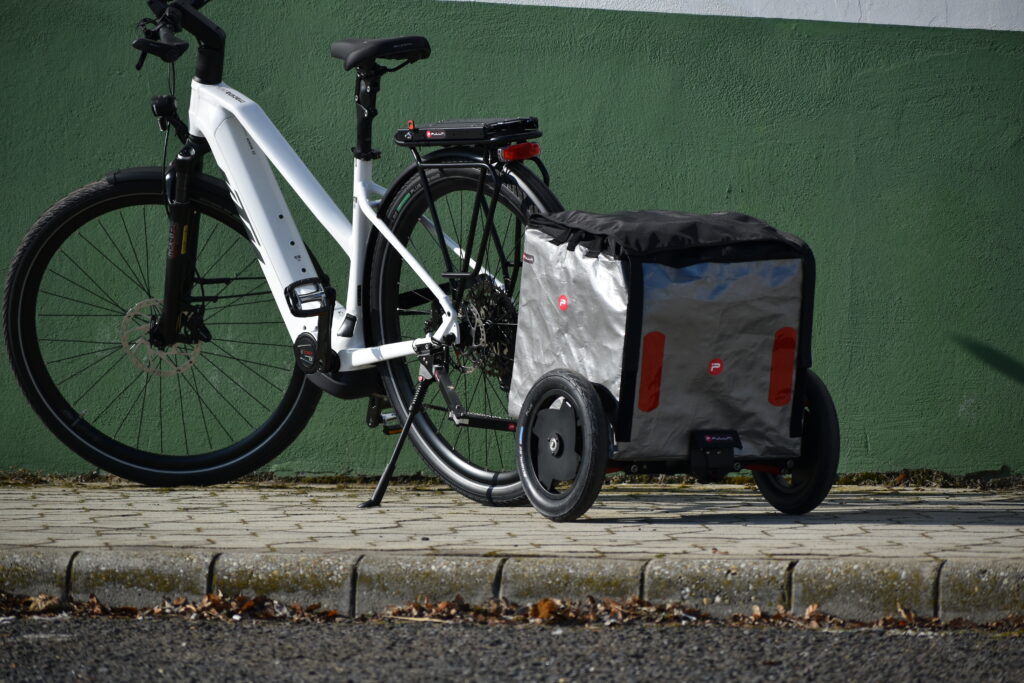 weißes KTM Pedelec Fahrrad mit Pullit Fahrradanhänger und silberner Tasche auf dem Gehweg vor grüner und weißer Wand