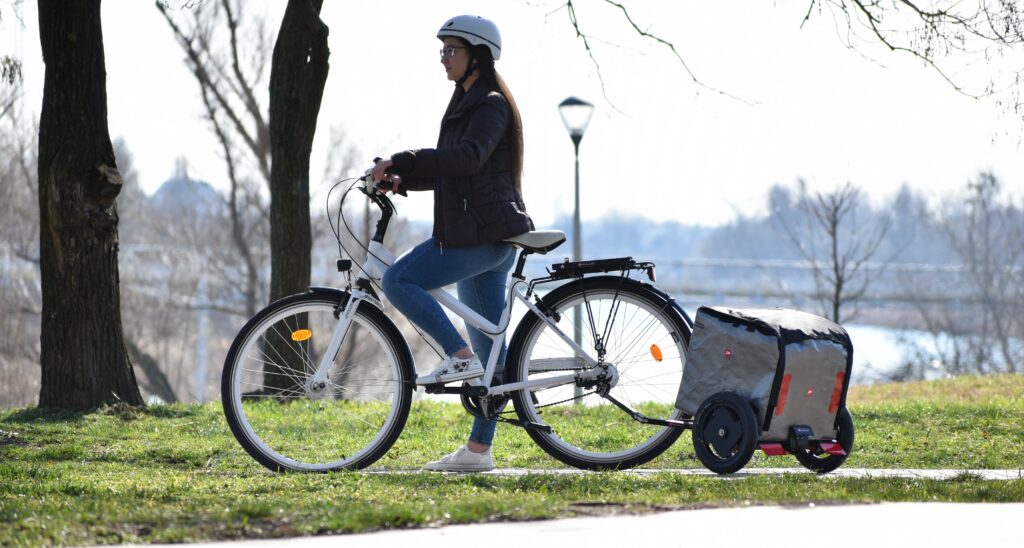 Junge Frau mit weißem elegantem Fahrrad und Pullit Fahrradanhänger vor Flusslandschaft.